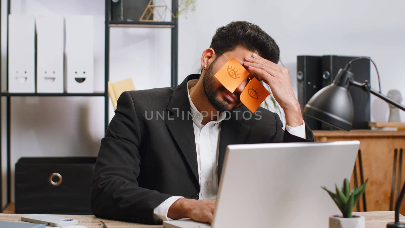 Inefficient tired young businessman working sleeping on laptop computer with eyes stickers on face at office workplace desk. Indian lazy manager freelancer man. Business people cheating to sleep