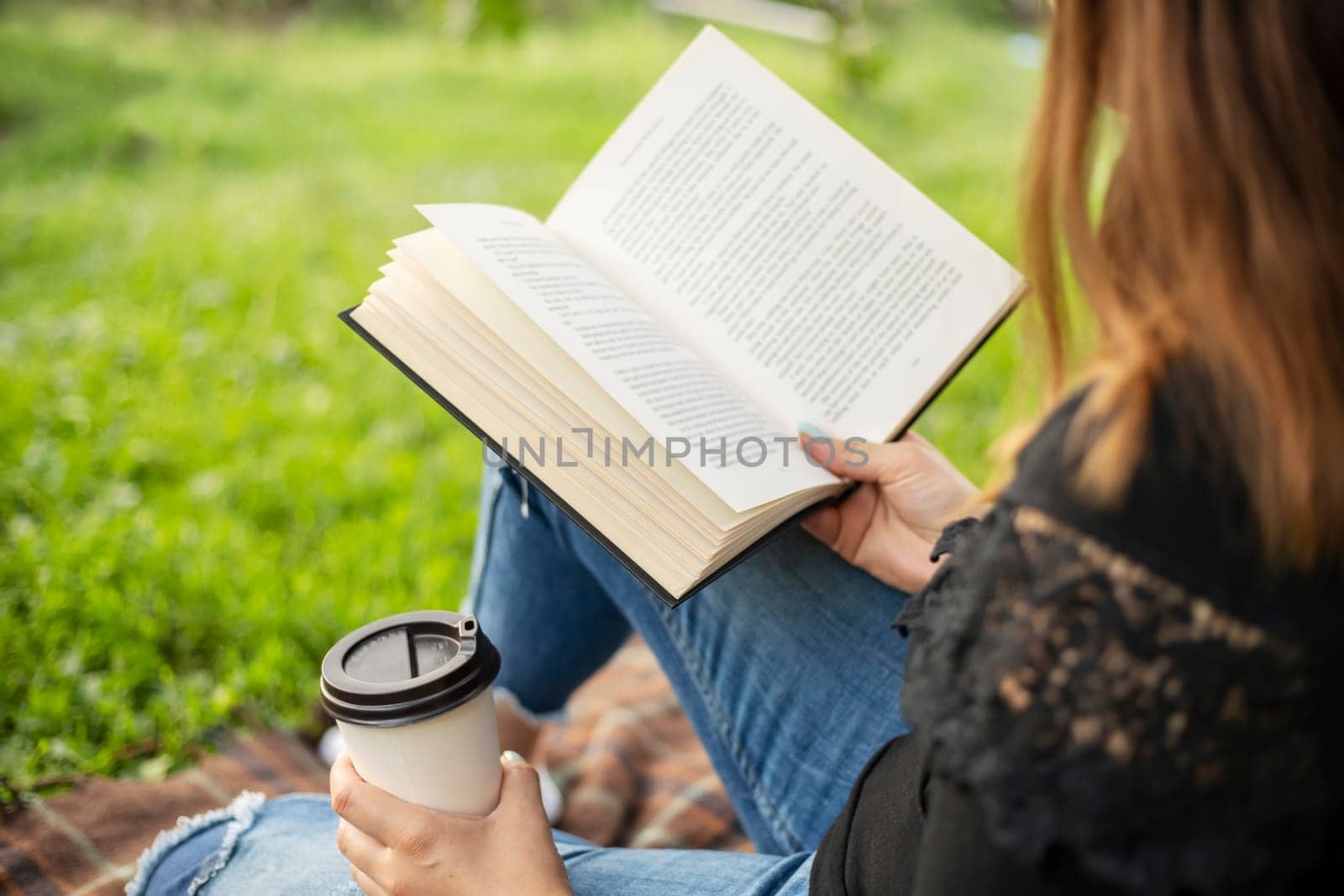 A woman sits near a tree in the park and holds a book and a cup with a hot drink in her hands. A woman in jeans and a t-shirt reading a book outdoor.