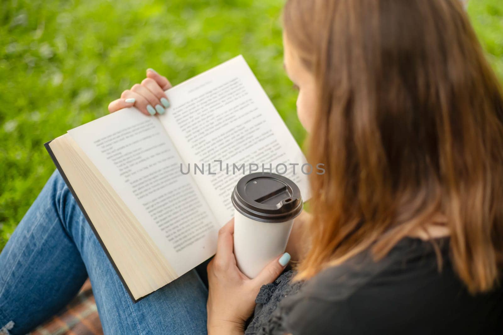 A woman sits near a tree in the park and holds a book and a cup with a hot drink in her hands. A woman in jeans and a t-shirt reading a book outdoor.