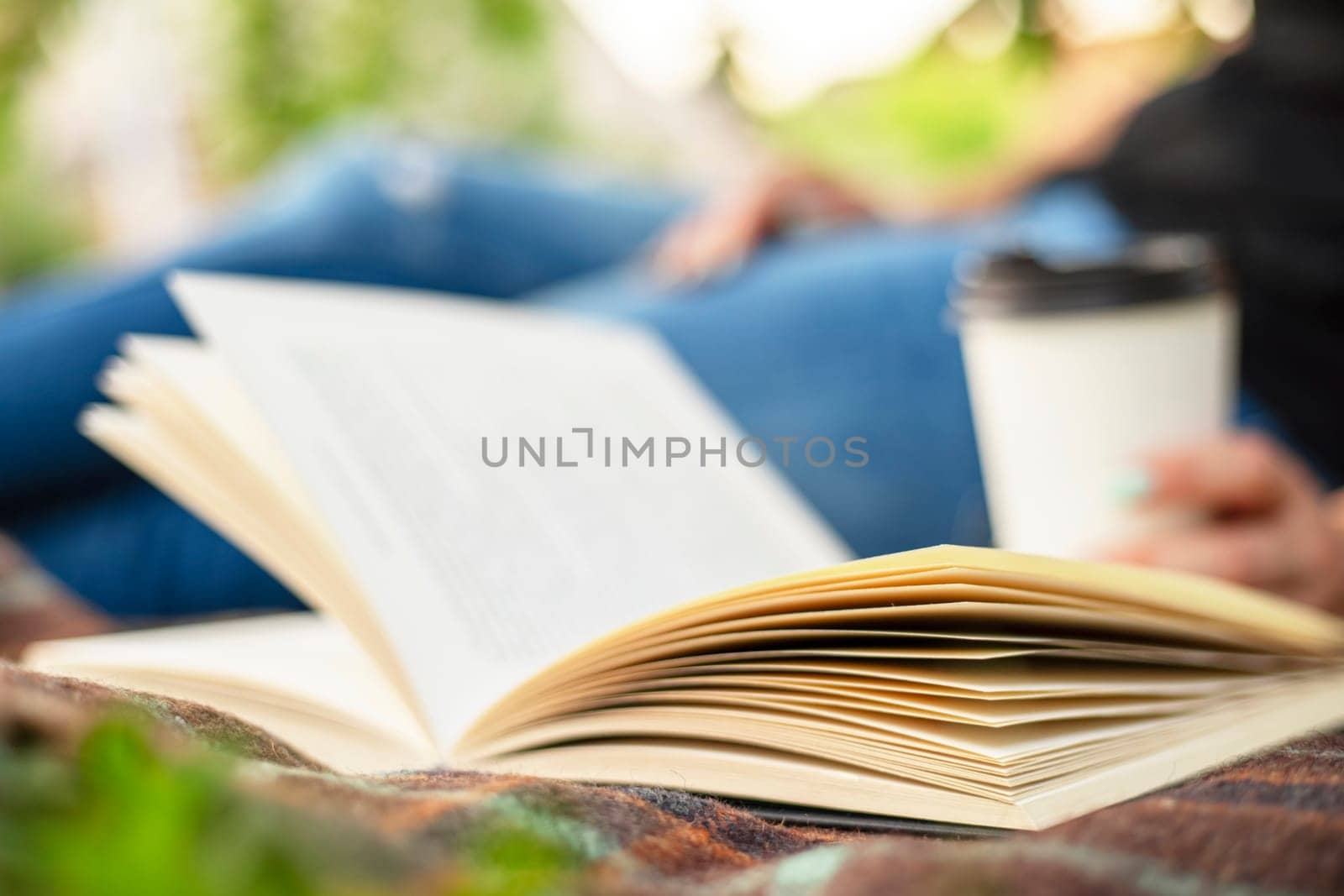 A woman in jeans with a book and a cup of coffee sits on a blanket in the park by andreyz