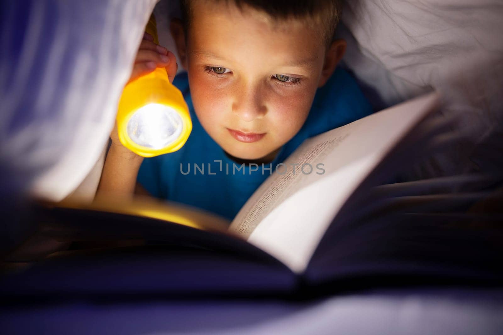 A young boy reading a book under the covers with a flashlight at dark night time.