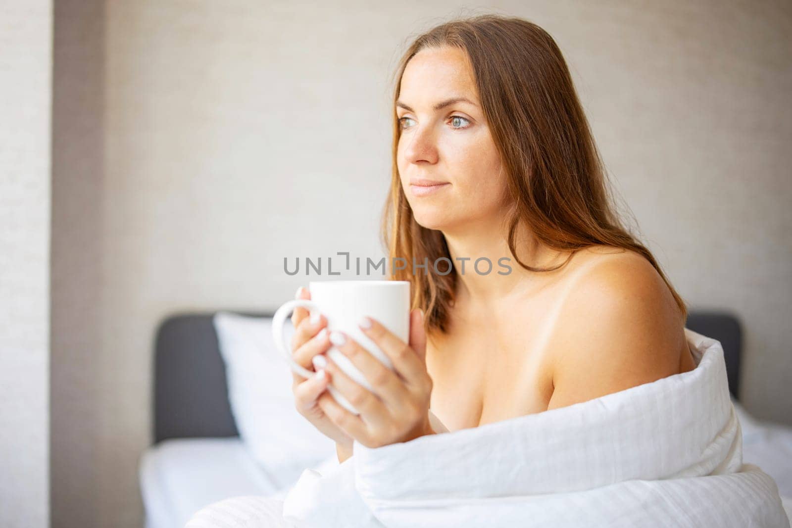 Beautiful young woman drinking a cup of coffee or tea while sitting on the bed after waking up in the morning. A woman is enjoying her drink in bed at home.