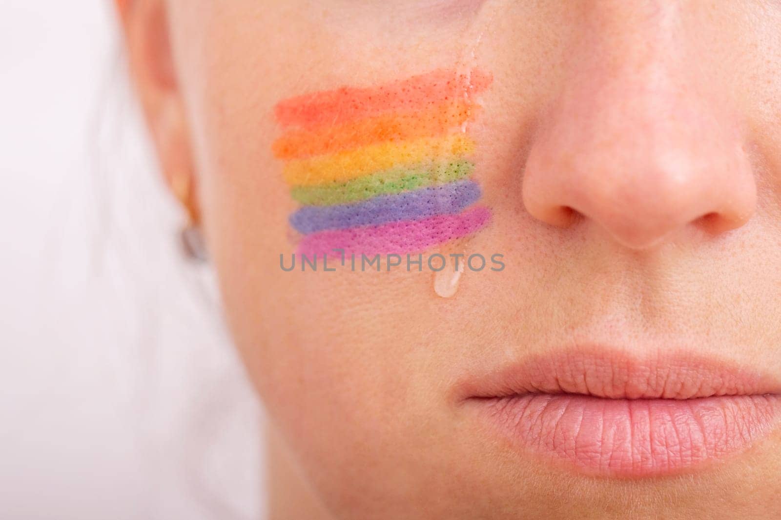 Part of the face of a young crying woman with a painted LGBT flag on her cheek. The concept of infringement of the rights and equality of LGBT people.