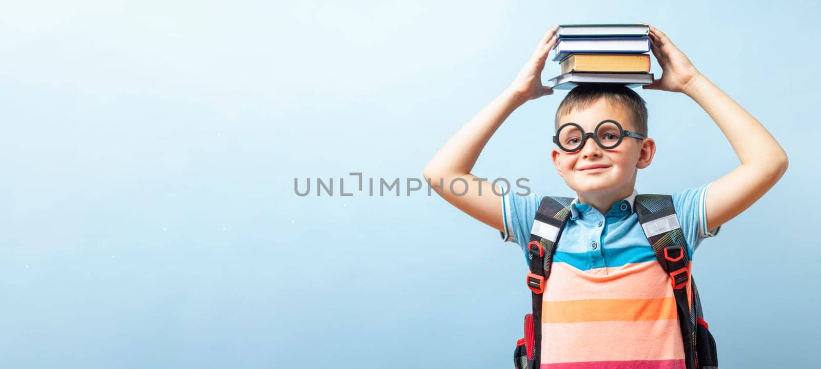 Funny smiling child school boy in glasses holding books on his head on blue background