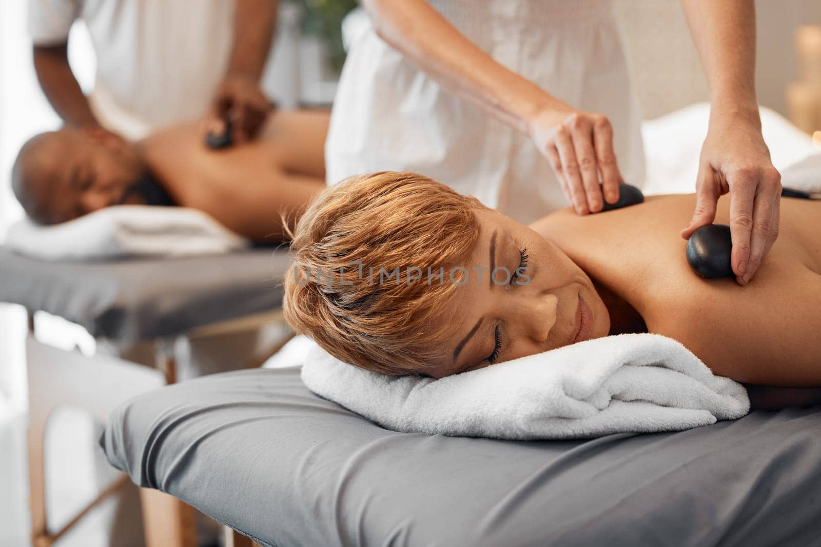 Spa, couple and massage for health and wellness while on bed for hot stone or rock therapy on back for zen, peace and relax. Man and woman customer in beauty salon for physical therapy and self care.