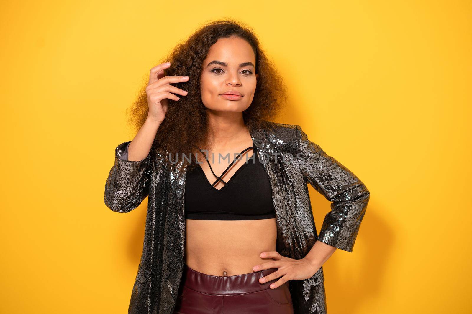 Posh beauty ethnic model girl with beautiful hairstyle smile looking at camera wearing shiny black jacket. Happy young girl fixing hair standing on yellow background. Studio shot.