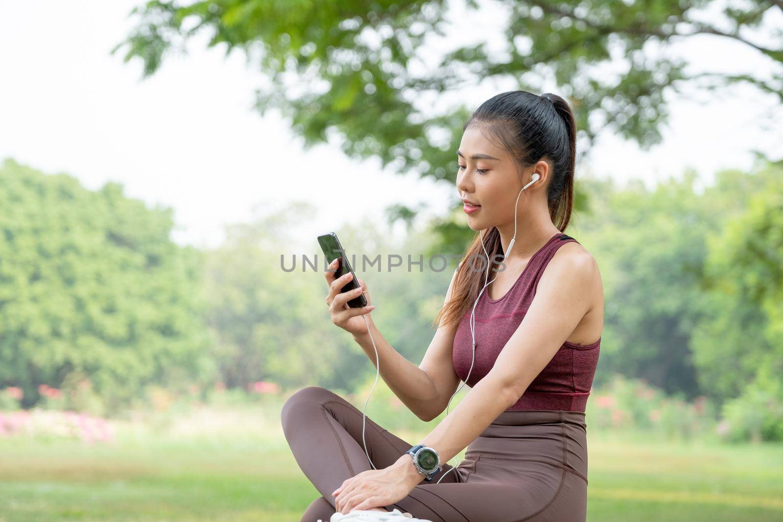 Asian beautiful woman with sportwear sit and relax also enjoy her mobile phone in park or garden in morning during exercise.