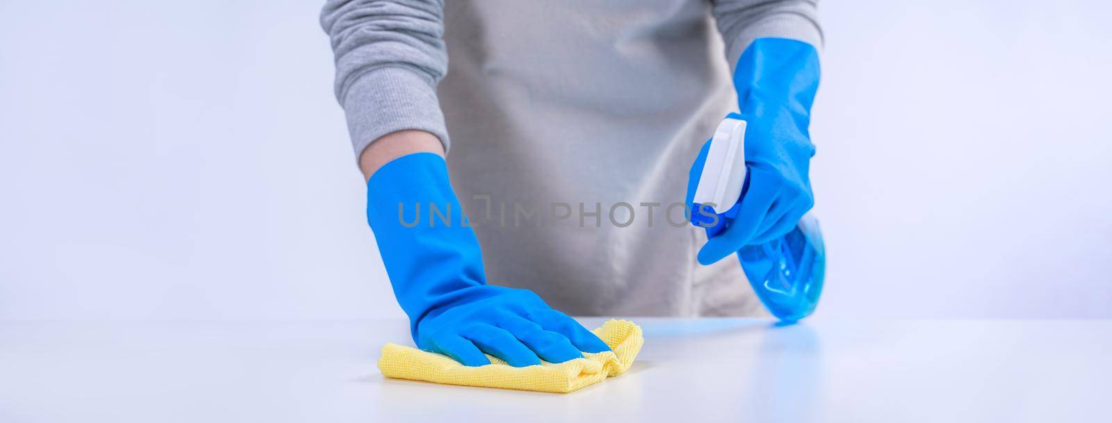 Young woman housekeeper in apron is cleaning, wiping down table surface with blue gloves, wet yellow rag, spraying bottle cleaner, closeup design concept. by ROMIXIMAGE