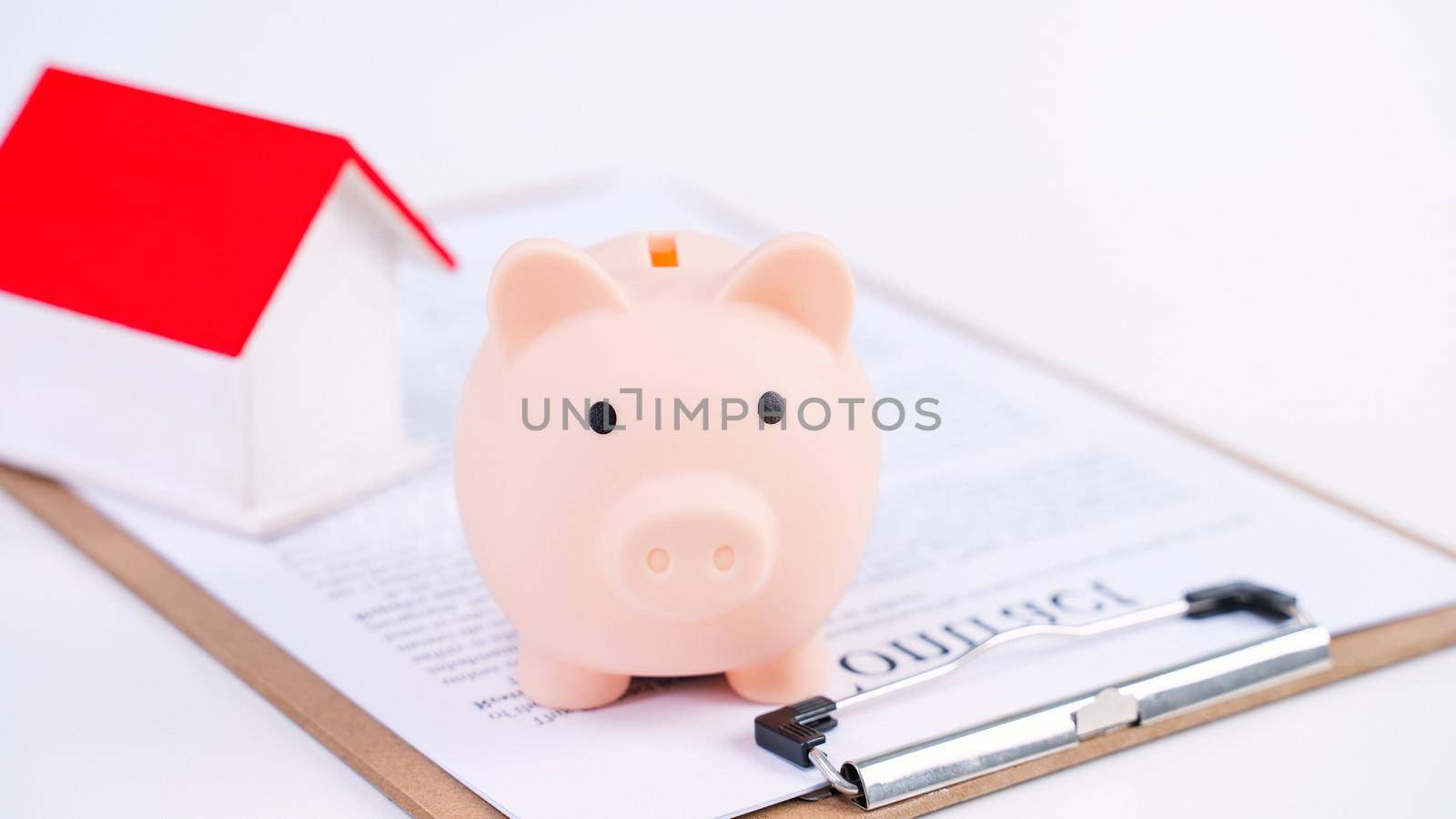 Piggy bank, beautiful wooden house model over a signed contract on white background, concept of saving money to buy a home insurance, close up, copy space. by ROMIXIMAGE