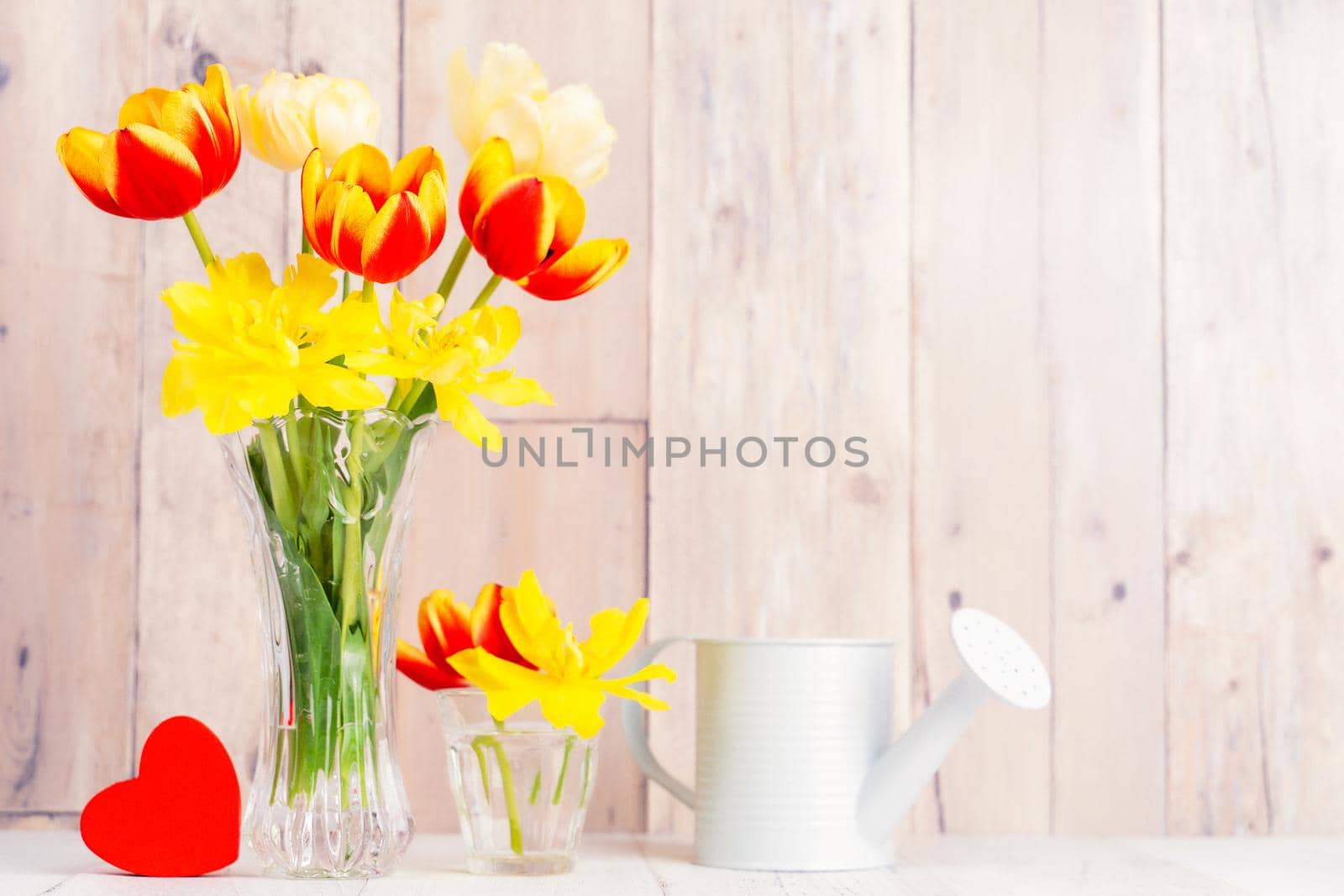 Tulip flower arrangement in glass vase with heart greeting, watering can decor on wooden table background wall, close up, Mother's Day design concept.