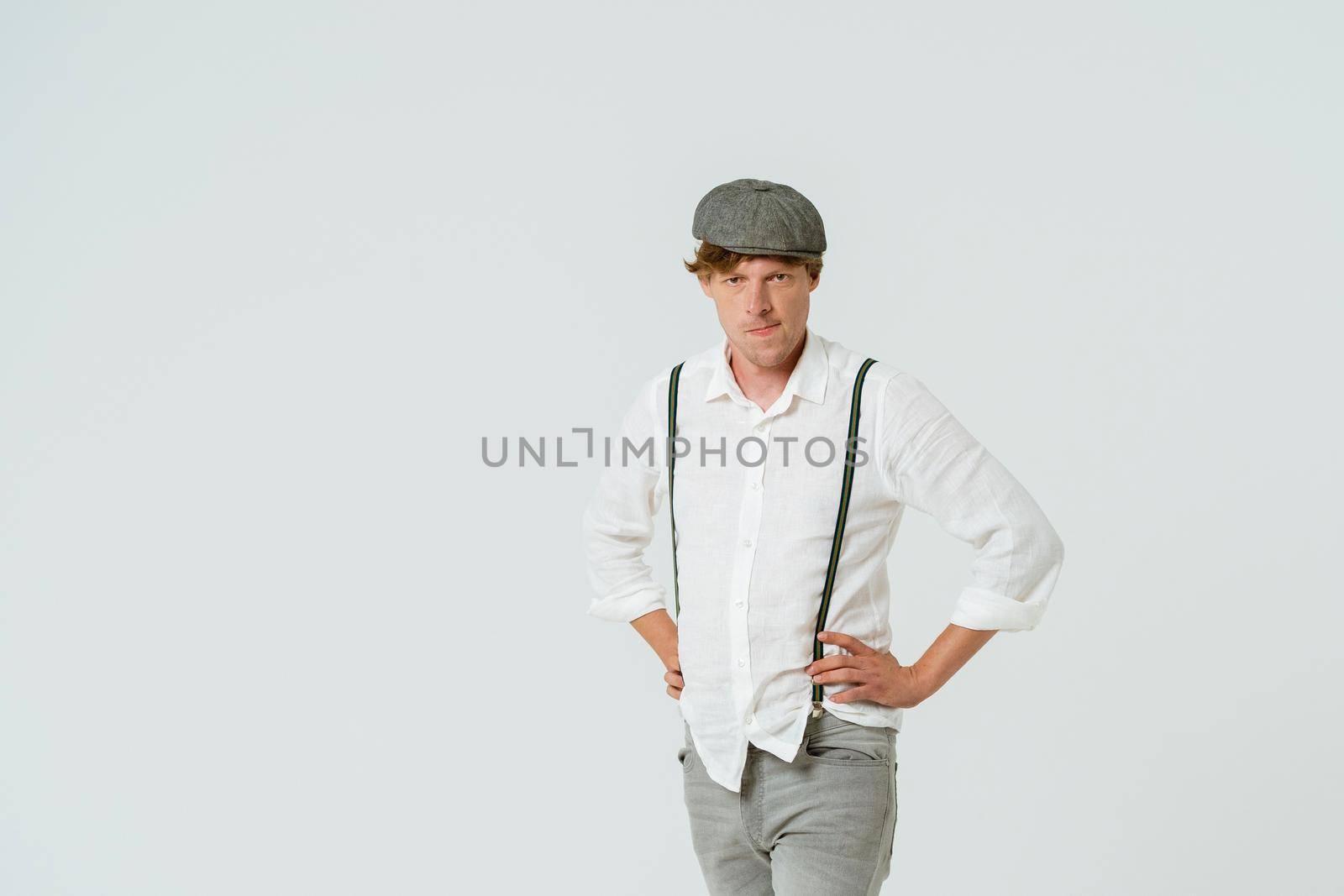 Handsome hipster man posing with hands on hips dressed in white shirt and grey jeans and wool cap isolated on white background. Copy space at left side by LipikStockMedia