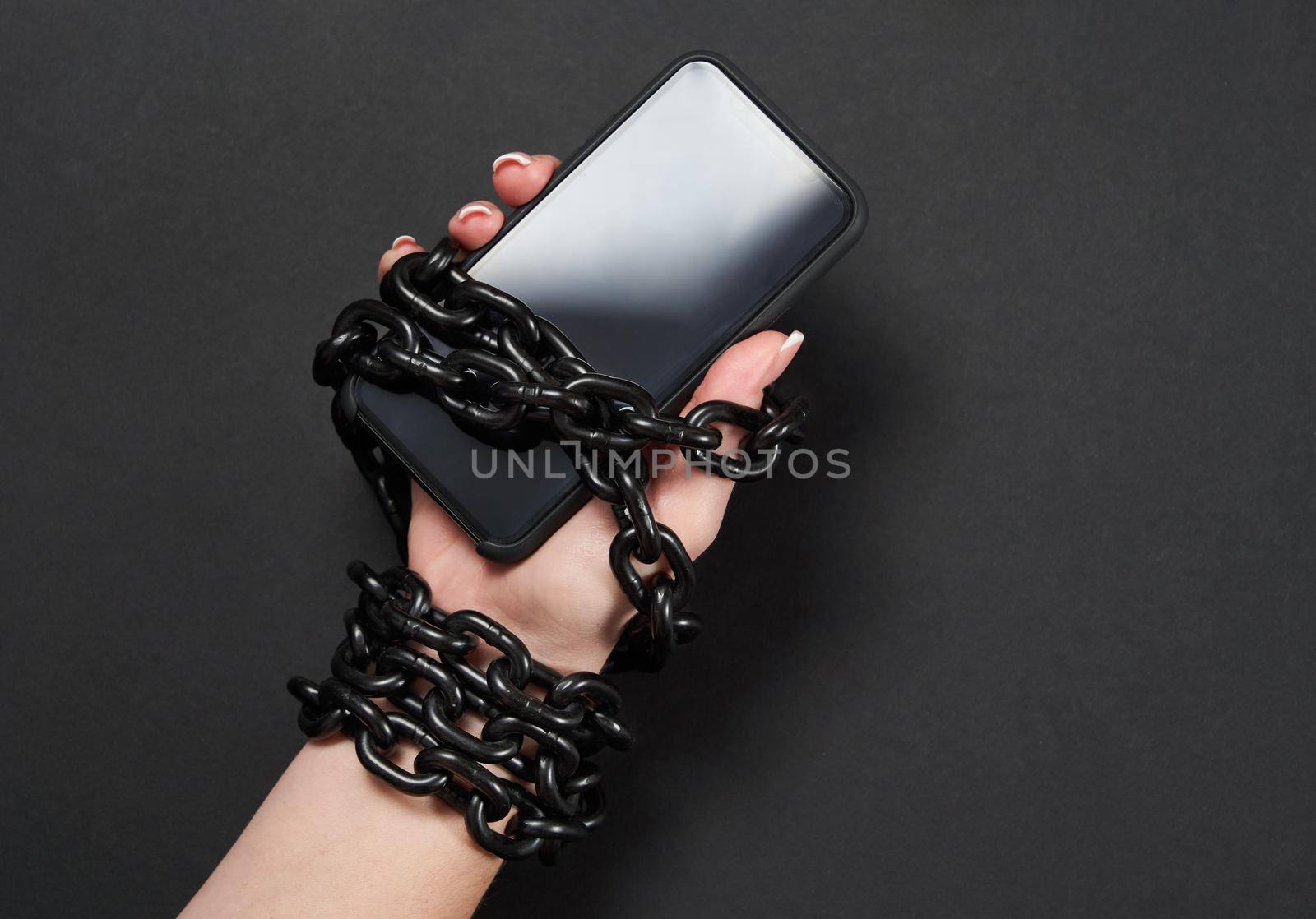 Iron chain that ties together hand and smartphone in concept of social media and internet addiction an black background by Mariakray