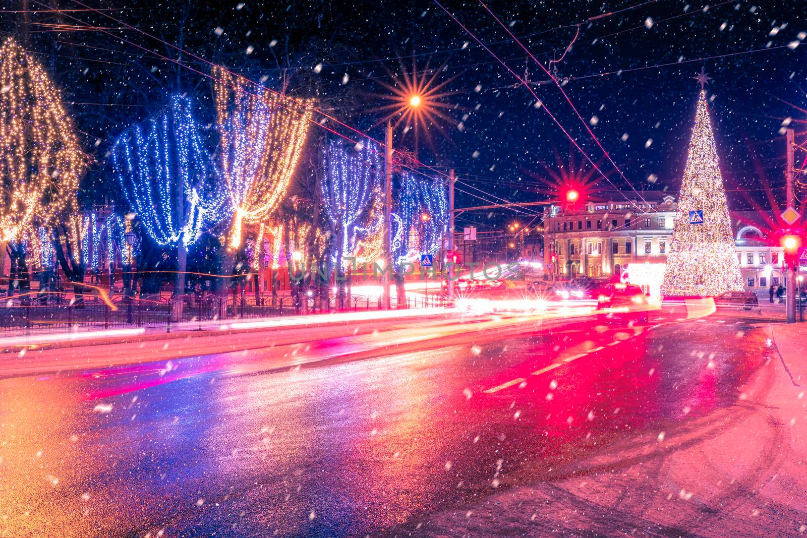 Night city with Christmas decorations, spruce and traces of headlights of moving cars, reflected in the wet road in a snowfall. by Eugene_Yemelyanov