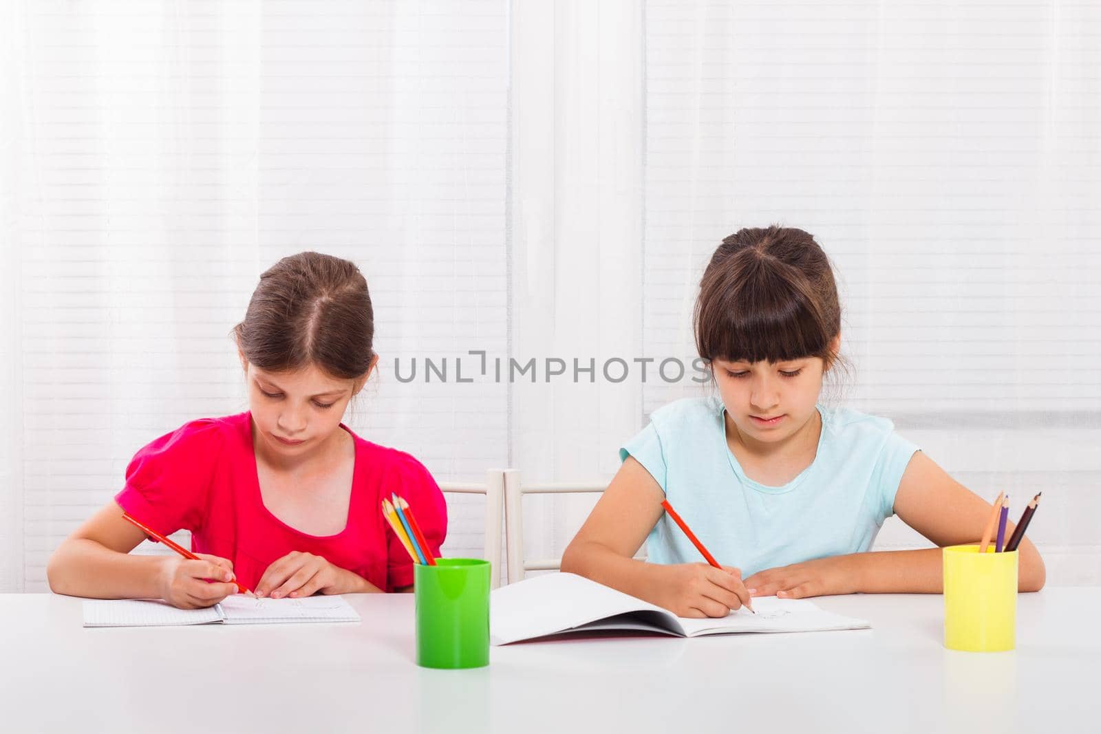 Two happy schoolgirls drawing at table in notebooks