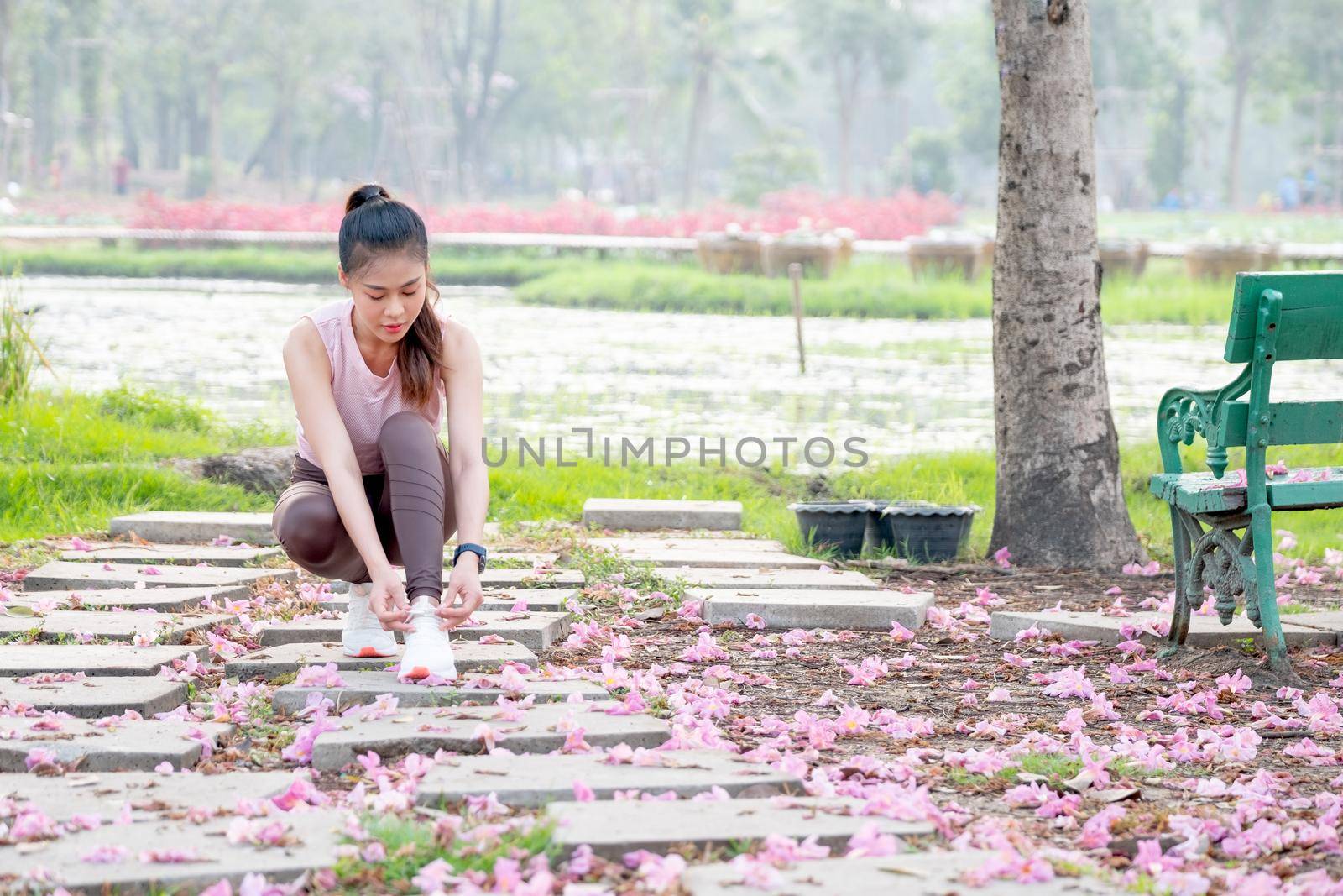 Wide shot of Asian sport woman tie shoelaces on walkway with flower on floor in part or garden during morning.