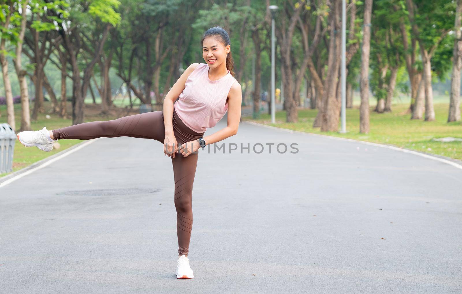 Asian sport woman stand with action of stretch right leg to side also look at camera and smile on road of park or garden with morning light.