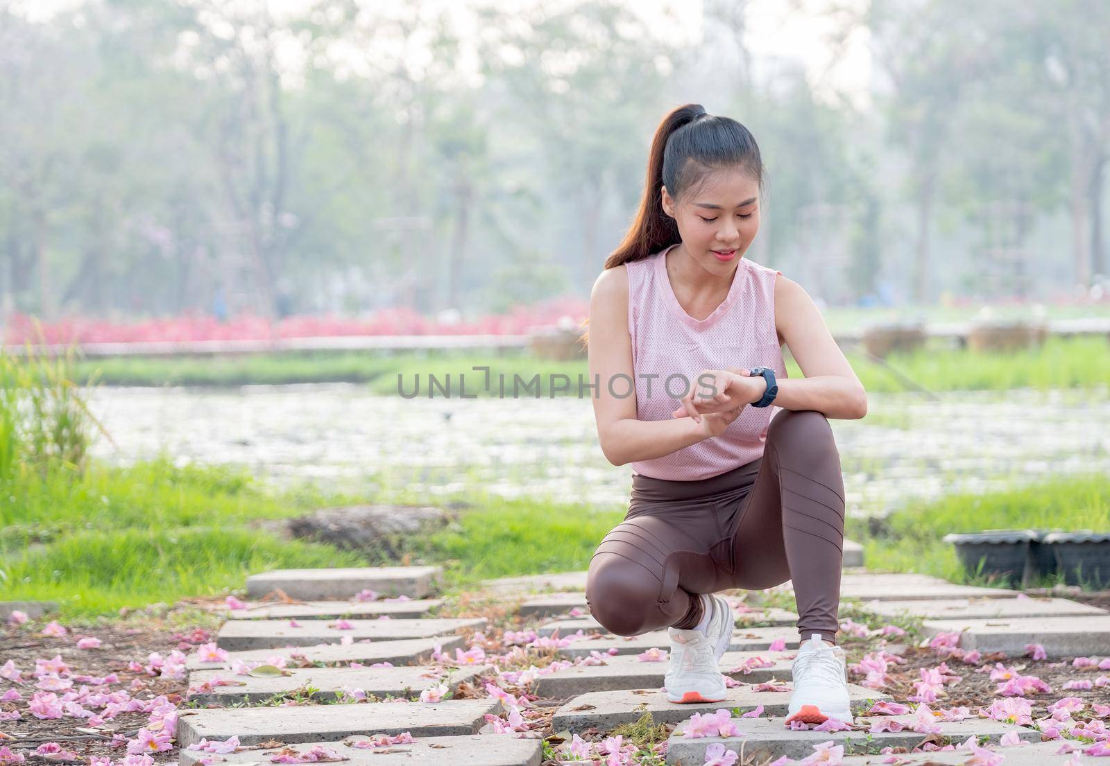 Asian woman with sportwear for jogging sit and look at her watch during exercise in garden or park in the morning with soft light.