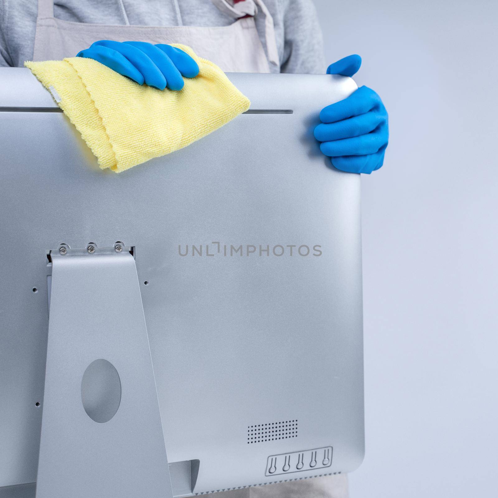 Young woman housekeeper in apron is doing cleaning silver computer screen with blue gloves, wet yellow rag, close up, copy space, blank design concept. by ROMIXIMAGE