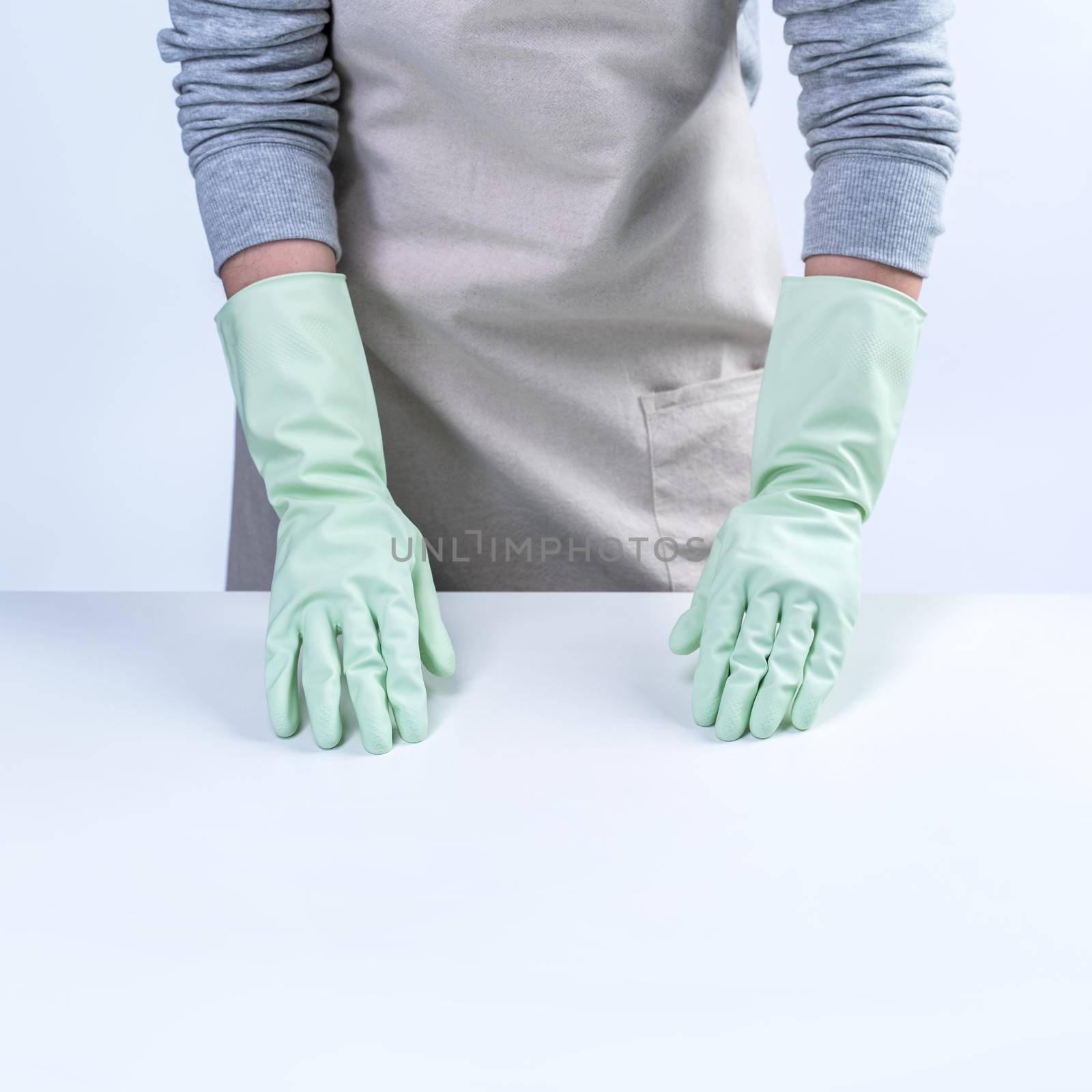 Young woman housekeeper in apron is wearing green gloves to clean the table, concept of preventing virus infection on white background, close up. by ROMIXIMAGE