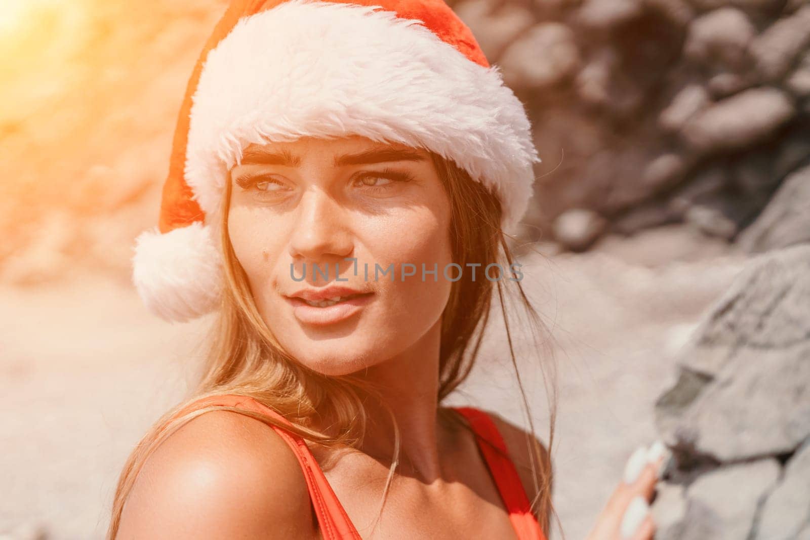 Woman summer travel sea. Happy tourist in red bikini and Santas hat enjoy taking picture outdoors for memories. Woman traveler posing on the beach surrounded by volcanic mountains, sharing travel joy by panophotograph