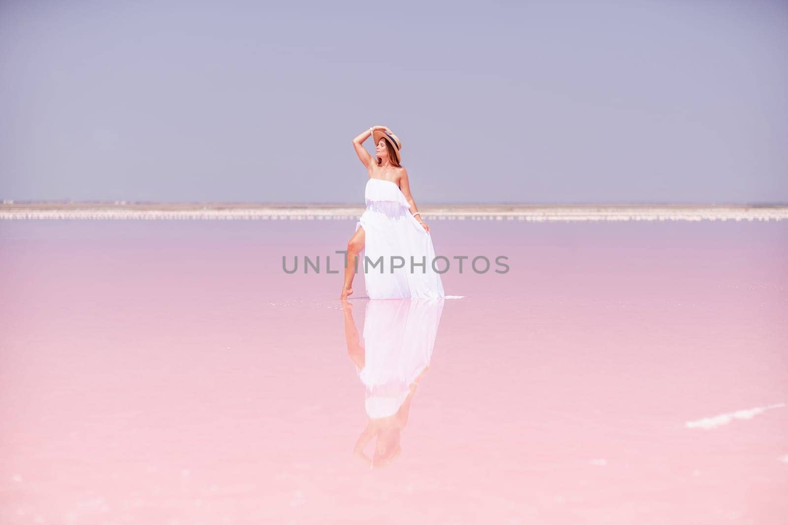 Woman in pink salt lake. She in a white dress and hat enjoys the scenic view of a pink salt lake as she walks along the white, salty shore, creating a lasting memory. by Matiunina