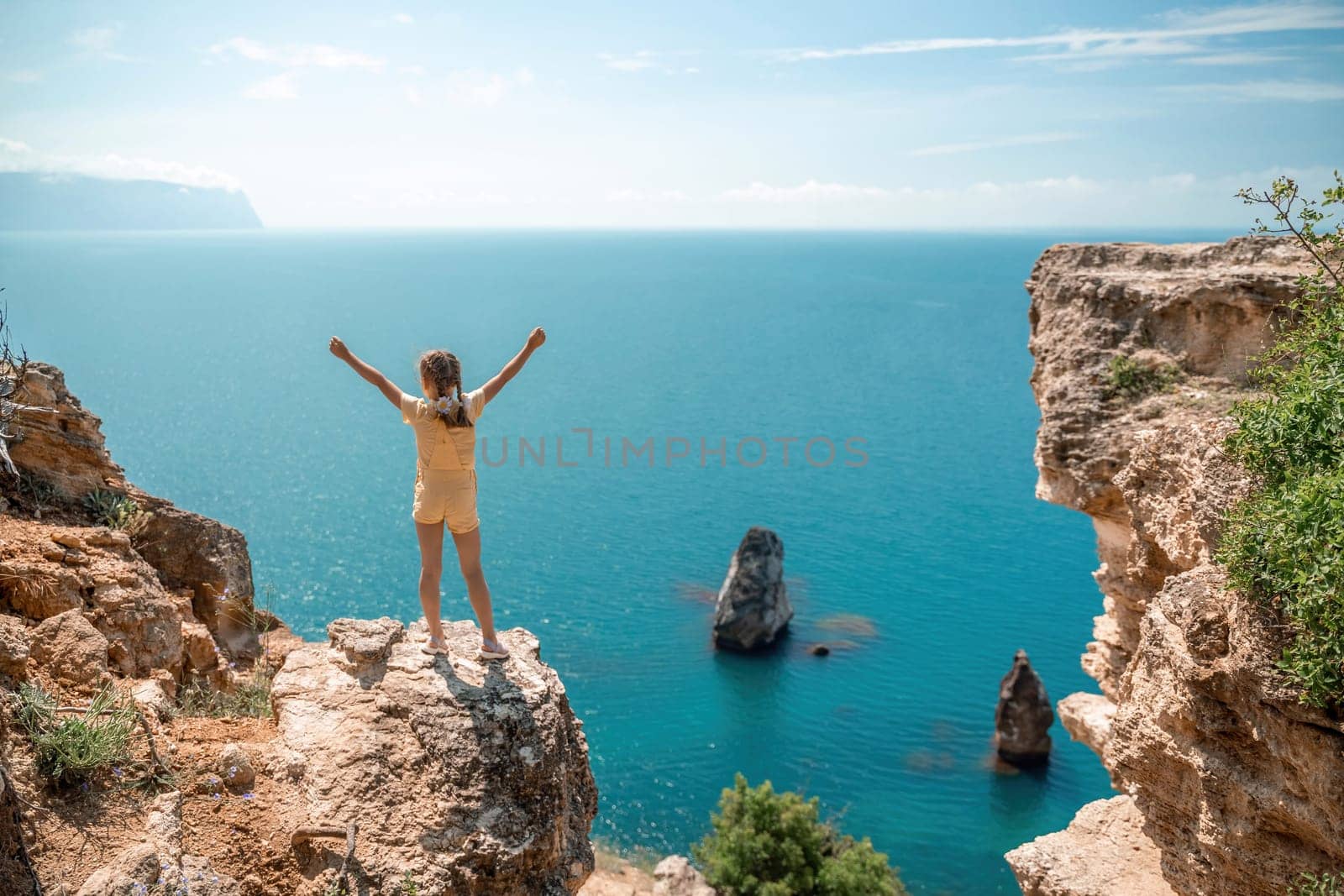 Happy girl stands on a rock high above the sea, wearing a yellow jumpsuit and sporting braided hair, depicting the idea of a summer vacation by the sea