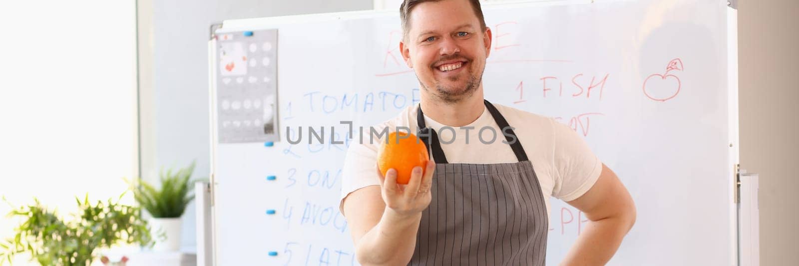 Male cook teacher in apron holds an orange and fresh vegetables and fruits on table by kuprevich