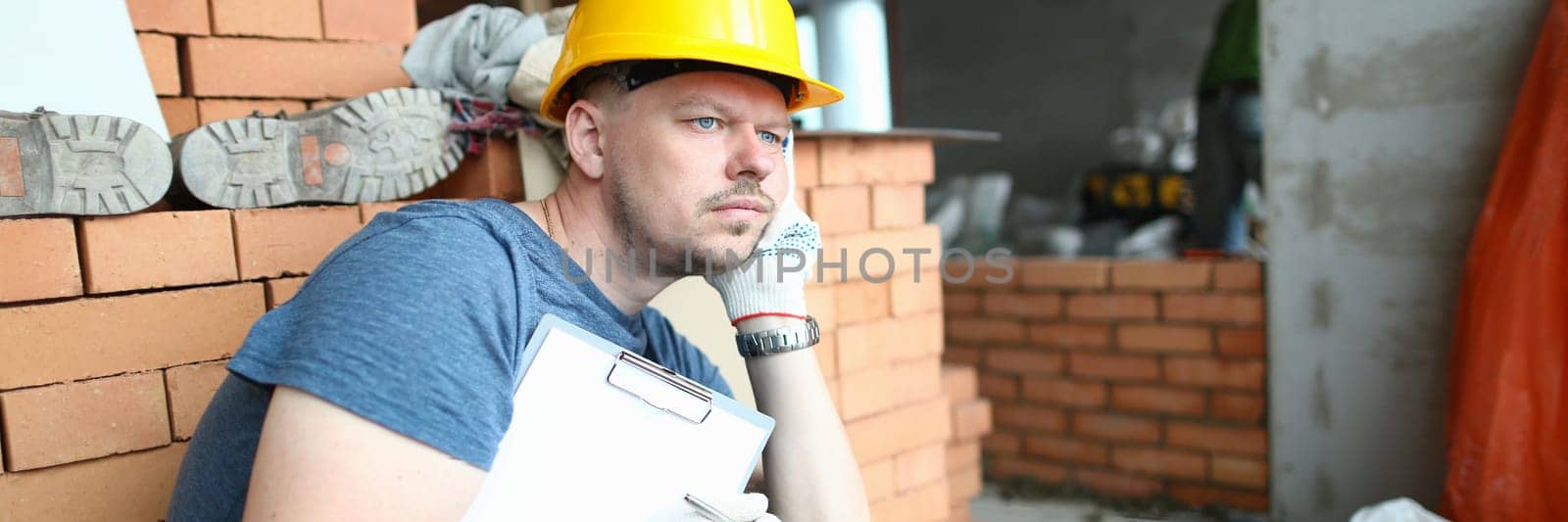 Thoughtful sad builder holding clipboard and looking into distance. Solving construction problem and builder depression