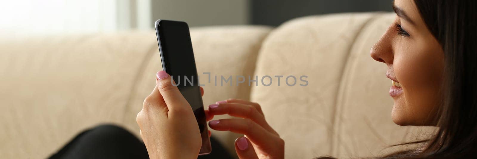Happy pleasant woman is resting on comfortable sofa holding smartphone in hands. Smiling young lady chatting on social networks and watching funny videos