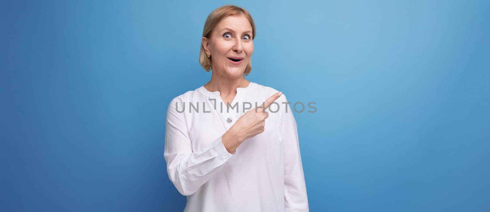 confident blond middle aged woman in white blouse pointing her finger to the side on studio background with copyspace.