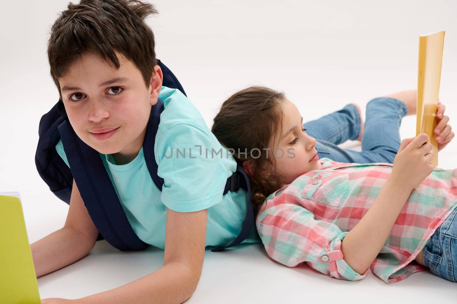 Portrait of Caucasian handsome preteen schoolboy with backpack and workbooks, lying near his younger sister, smiling looking at camera, isolated on white studio background. School kids. Back to school