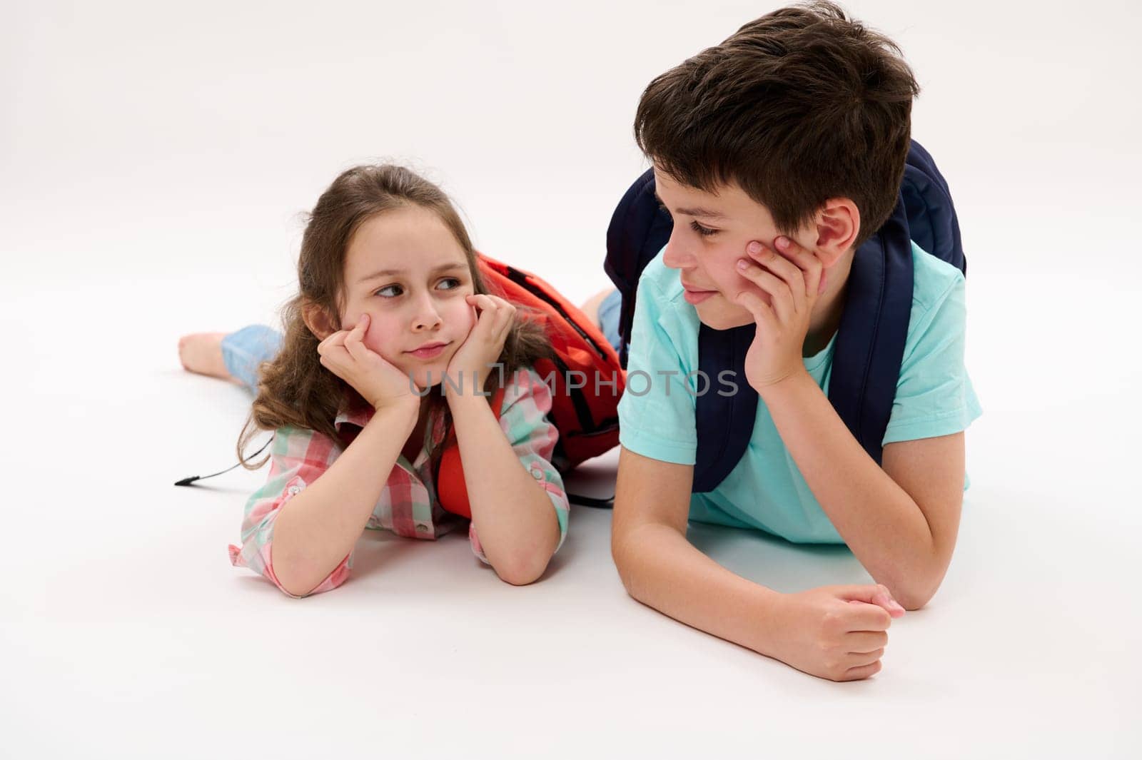 Two Caucasian adorable happy smart school kids, a teen boy a little girl with backpacks talking together during recreation at school, smiling, lying on their belly, isolated on white studio background