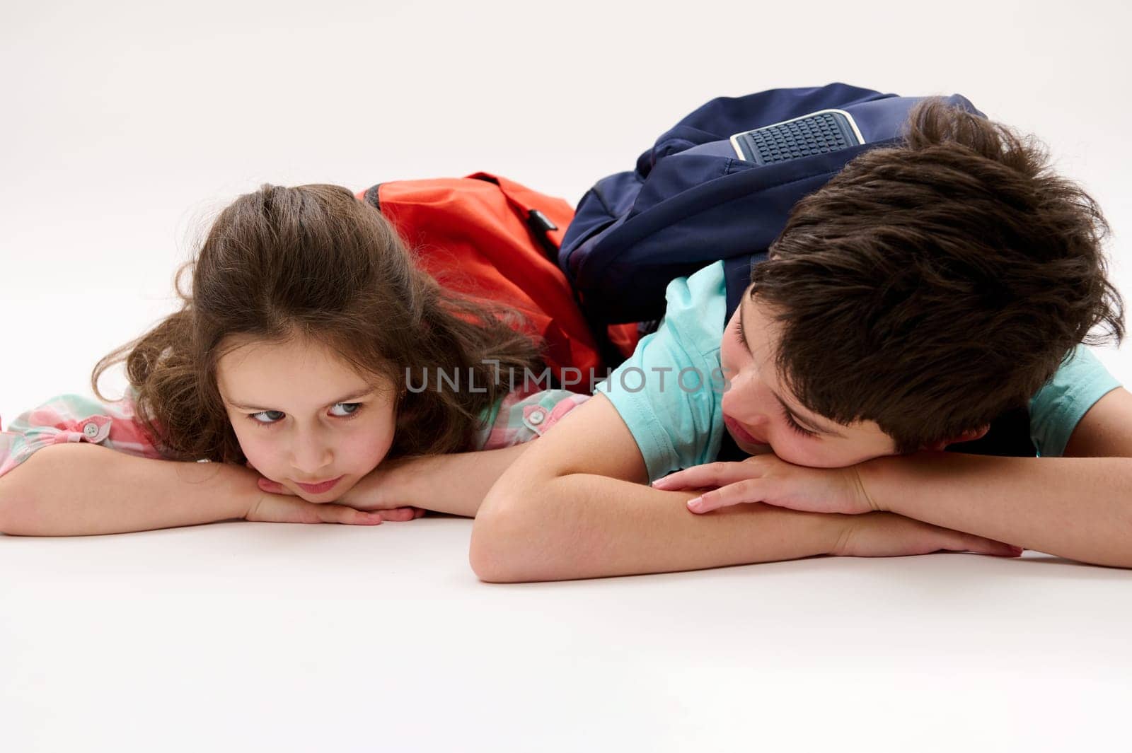 Conceptual emotional portrait of a happy little preschool girl and her brother, preteen school boy with backpacks, talking to each other in school recreation time, lying on belly over white background