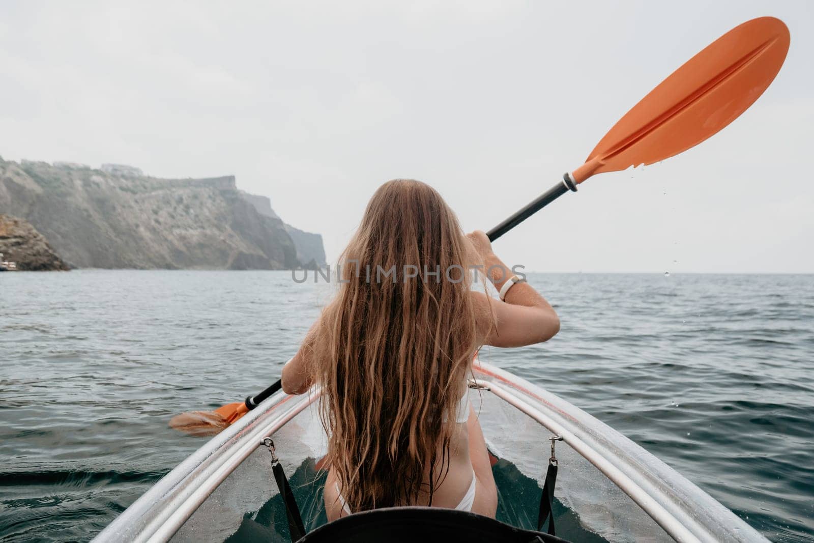 Woman in kayak back view. Happy young woman with long hair floating in transparent kayak on the crystal clear sea. Summer holiday vacation and cheerful female people relaxing having fun on the boat by panophotograph