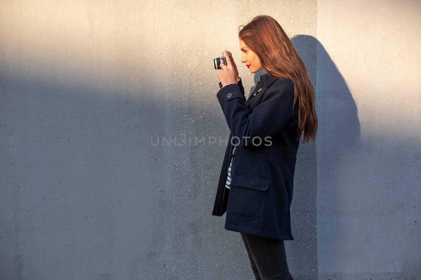 Smiling female photographer in jacket standing in front of wall ready to make new photo. Adorable young brunette woman in trendy outfit posing on concrete wall background with camera