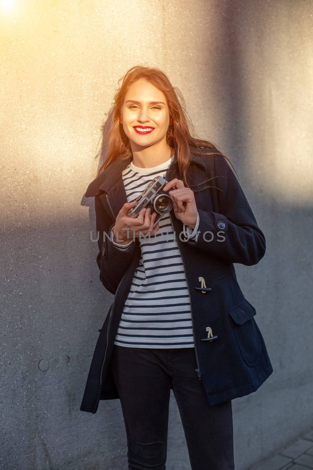 Smiling female photographer in jacket standing in front of wall ready to make new photo. Adorable young brunette woman in trendy outfit posing on concrete wall background with camera