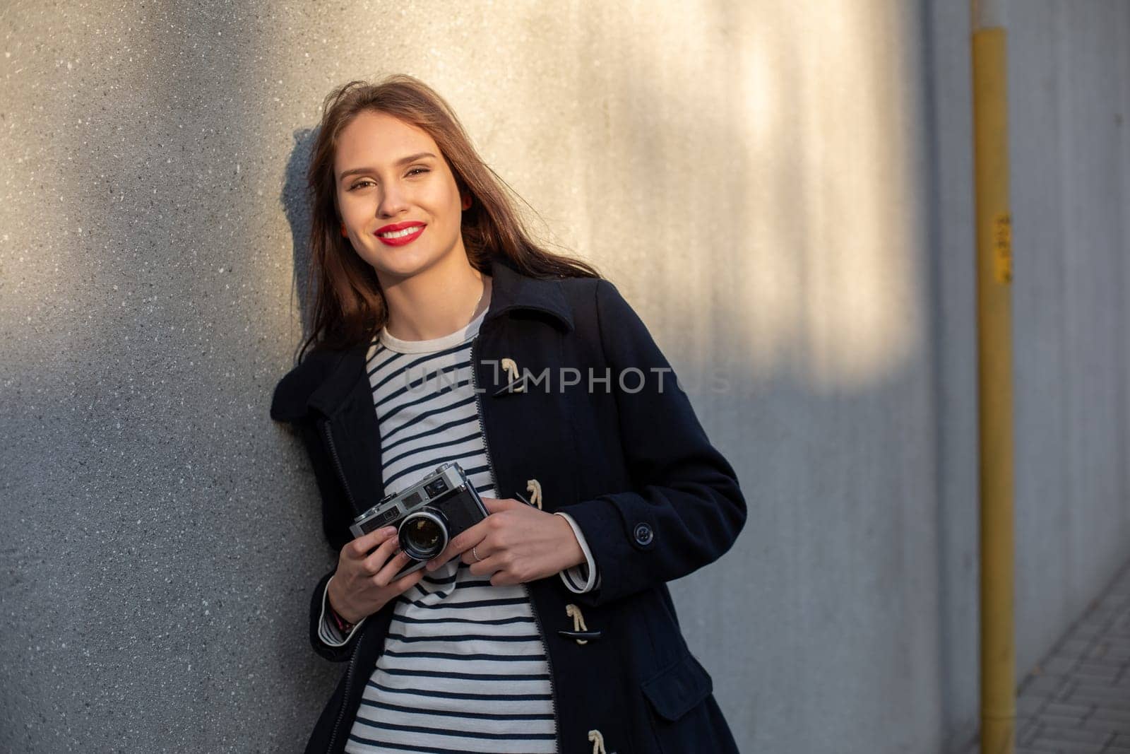 Smiling female photographer in jacket standing in front of wall ready to make new photo. Adorable young brunette woman in trendy outfit posing on concrete wall background with camera