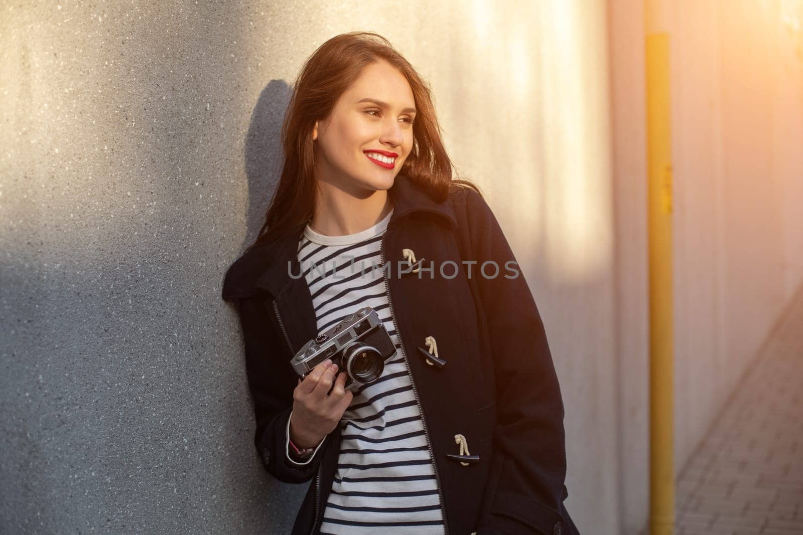 Smiling female photographer in jacket standing in front of wall ready to make new photo. Adorable young brunette woman in trendy outfit posing on concrete wall background with camera