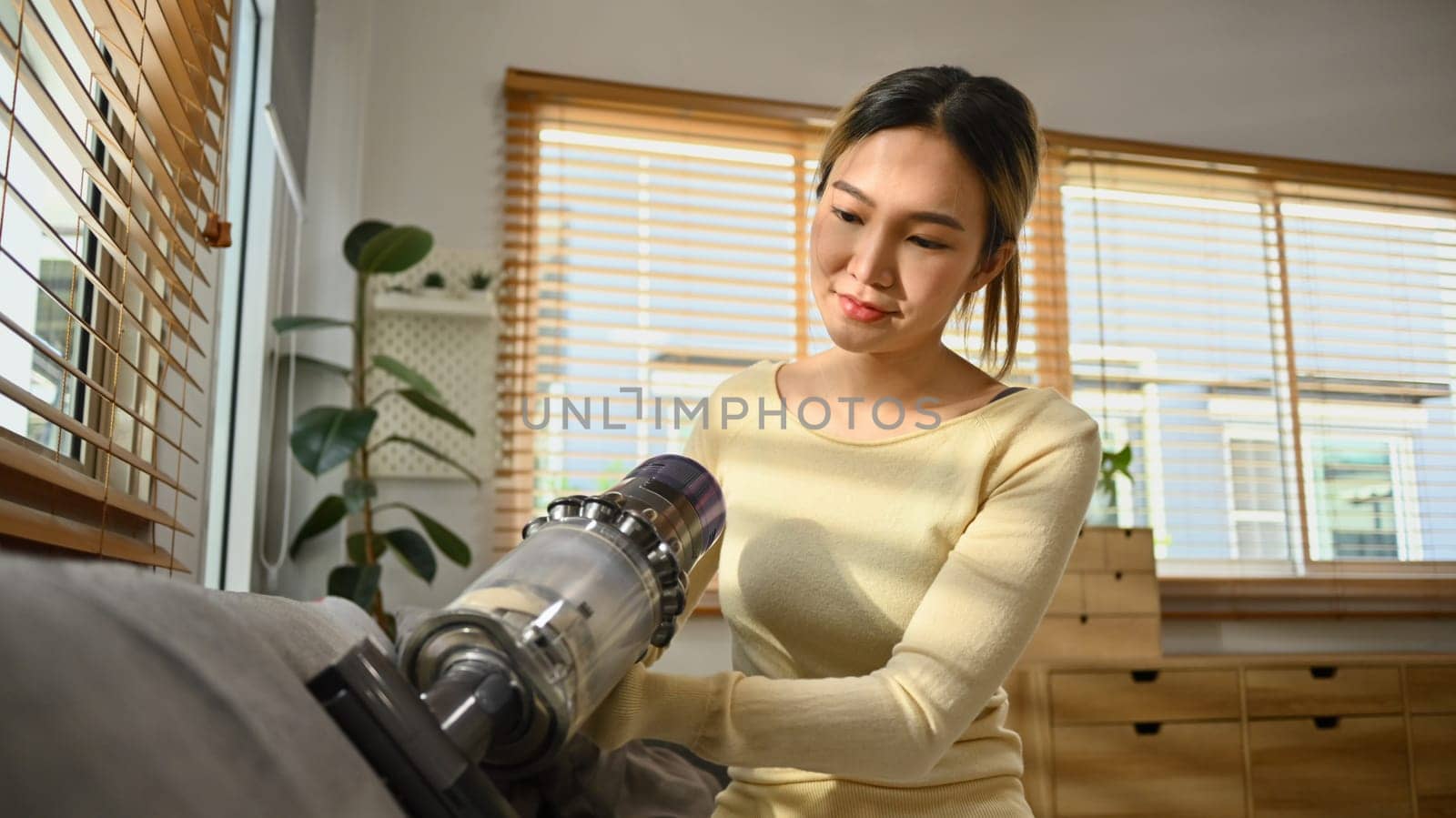 Beautiful female housewife vacuuming sofa with cordless handheld vacuum cleaner. Housework concept.