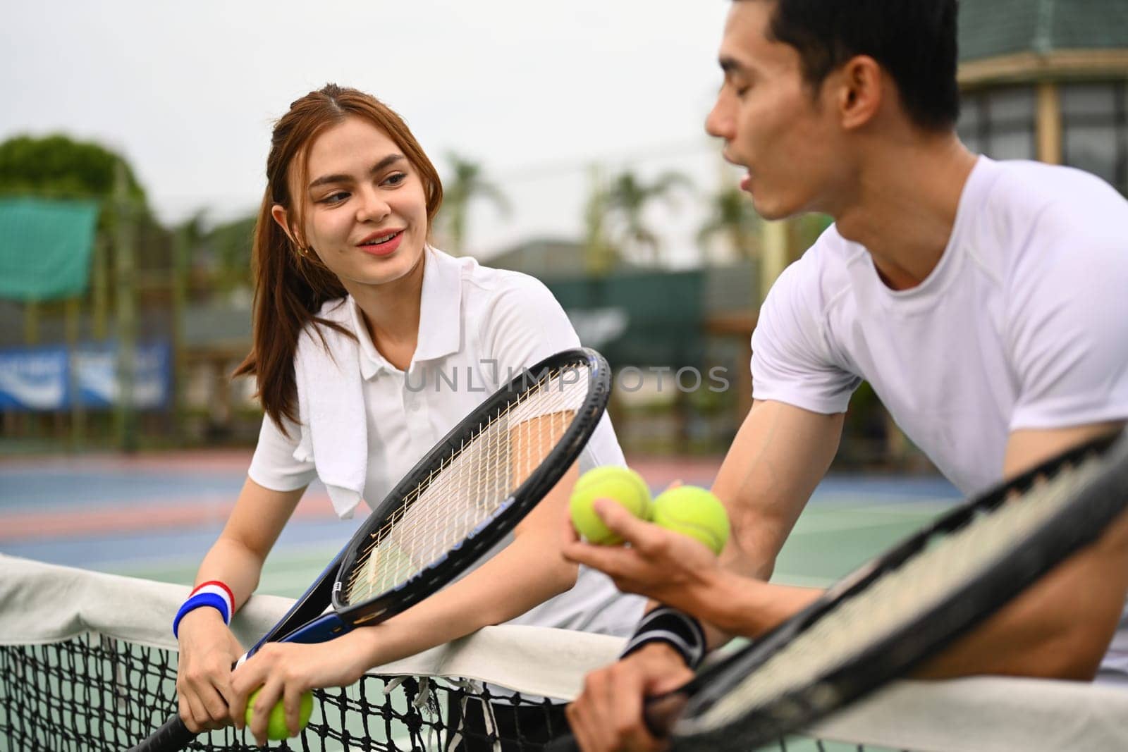 Smiling sportswoman holding racket and talking with male tennis coach, standing near net on the court.