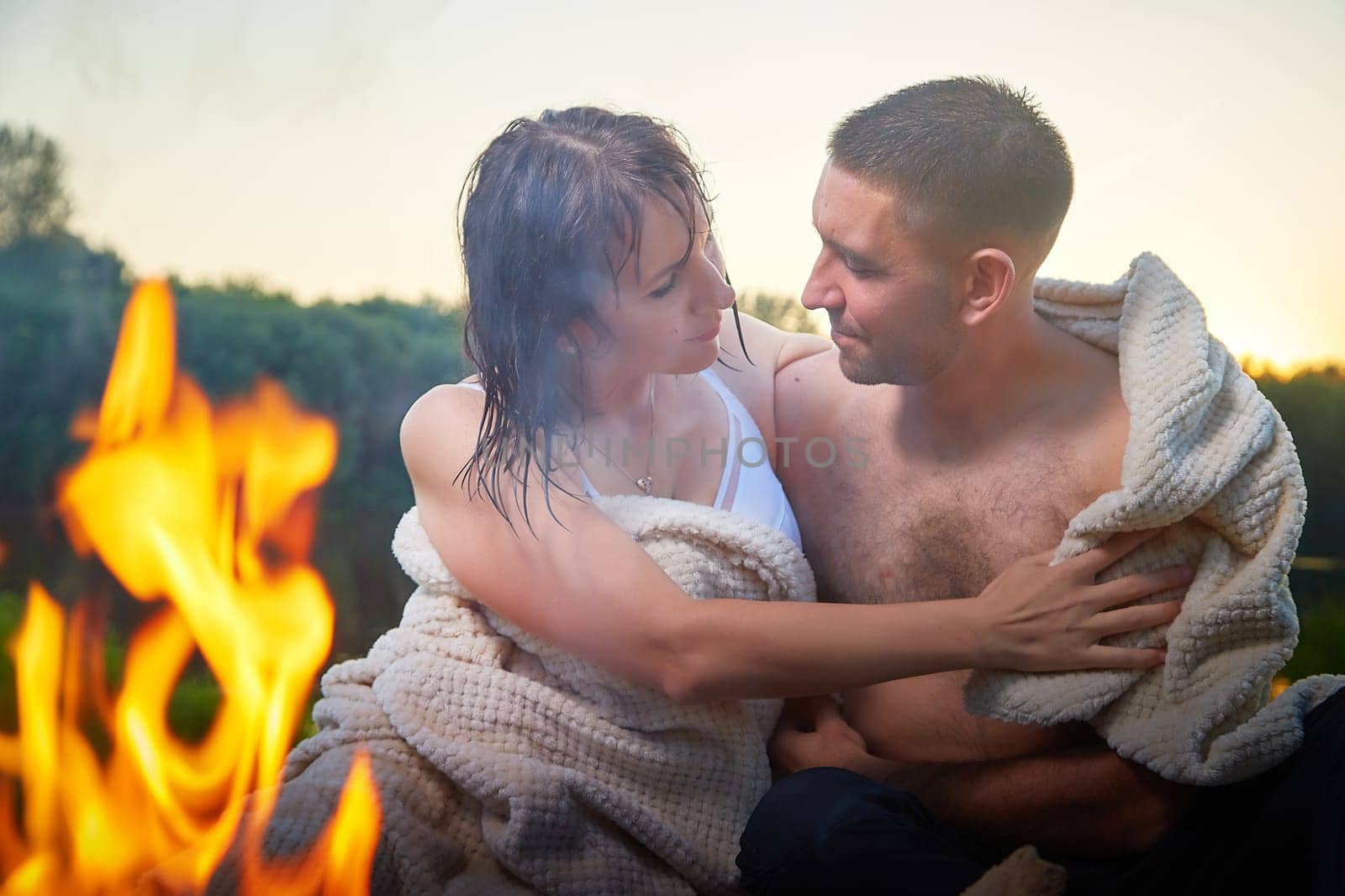 Happy Couple relaxin, having fun and hugs with fire in camping on nature near water of river or lake in summer sunny evening in sunset. Family or lovers have date and rest outdoor. Concept of love