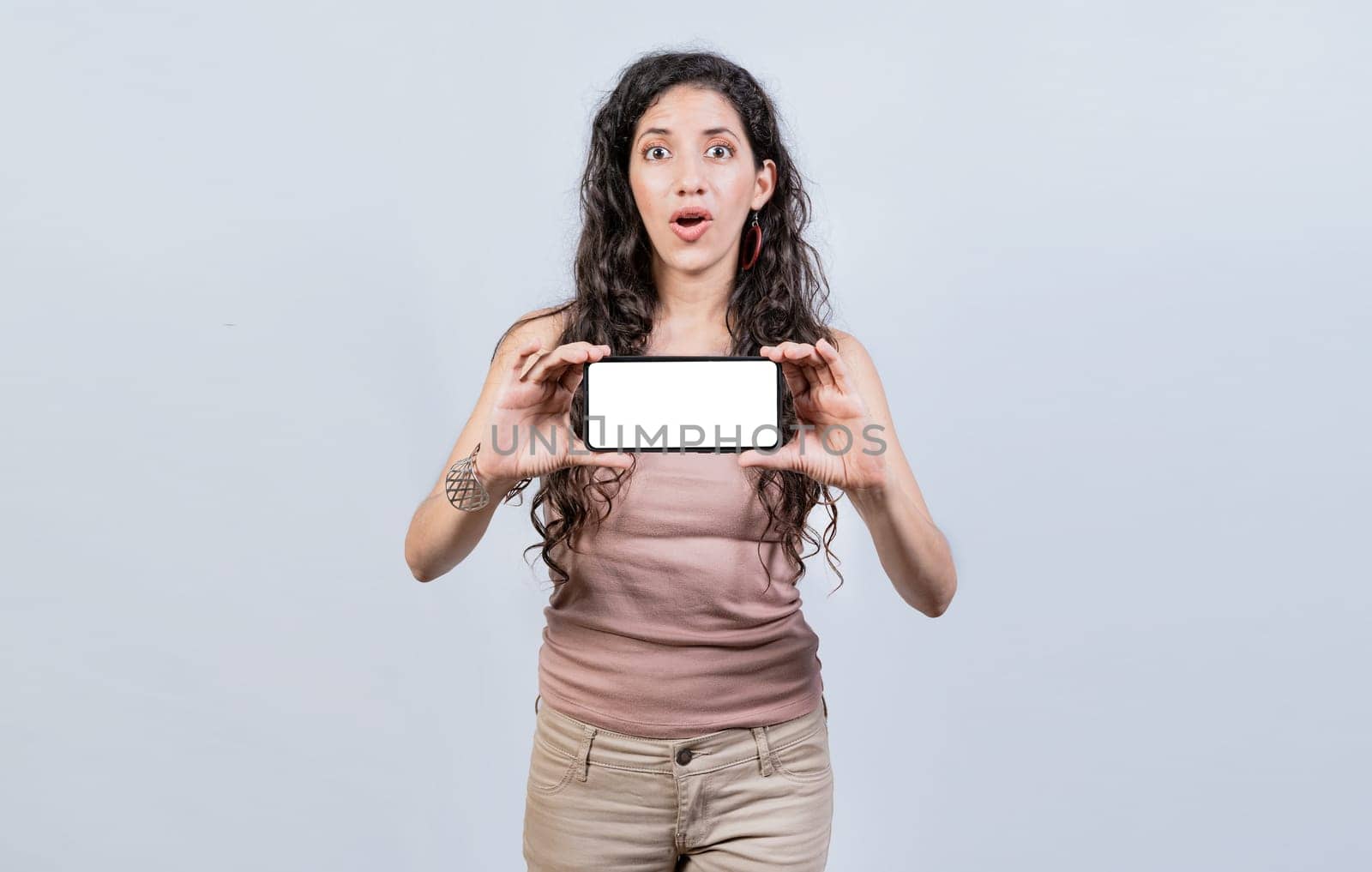 Surprised girl showing blank screen of smart phone. Latin girl showing horizontal screen of cell phone, Amazed young woman showing ad on cellphone by isaiphoto