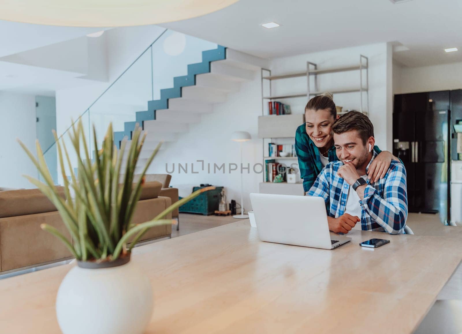 A young married couple is talking to parents, family and friends on a video call via a laptop while sitting in the living room of their modern house.