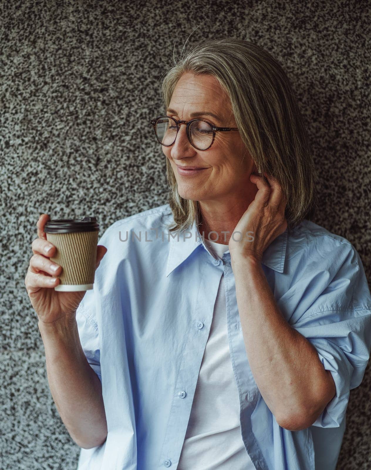 Coffee culture concept. Senior old woman enjoying paper cup of coffee on urban background of marble wall. Woman is depicted savoring moment, taking moment of relaxation and enjoyment with beverage. High quality photo