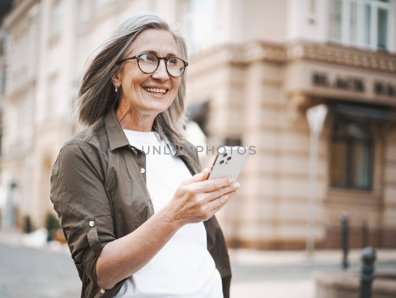 Old caucasian woman texting message on mobile phone while standing on street in old town of Europe. Seamless blend of modern technology with historic charm of surroundings. . High quality photo