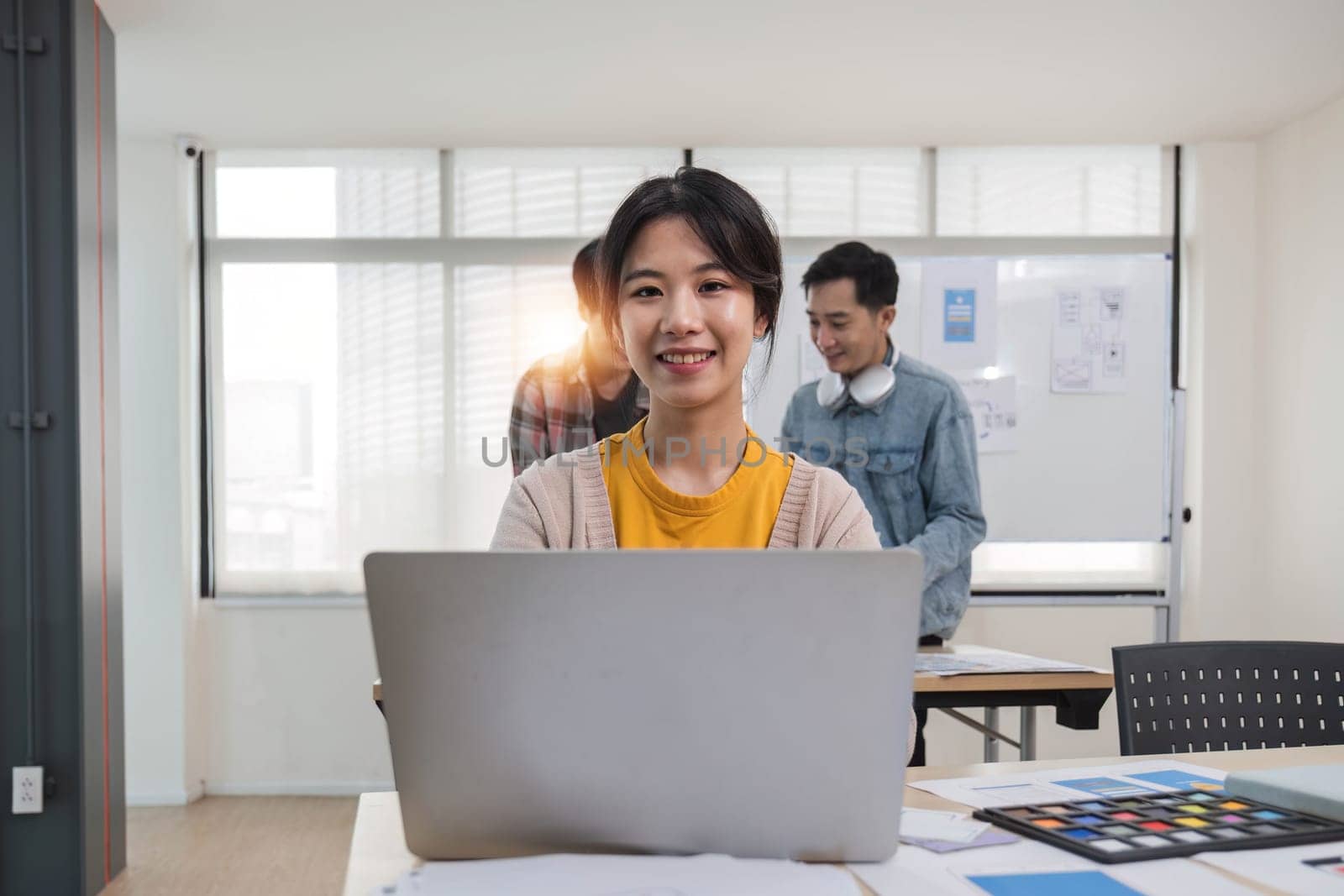 A focused young Asian female marketing assistant is working on her work on her laptop at her desk while her coworkers are working in the room. by wichayada