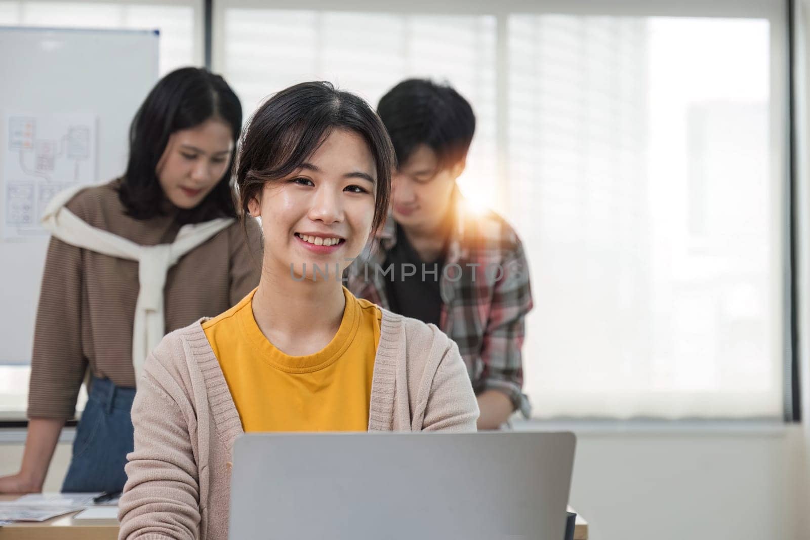 A focused young Asian female marketing assistant is working on her work on her laptop at her desk while her coworkers are working in the room. by wichayada