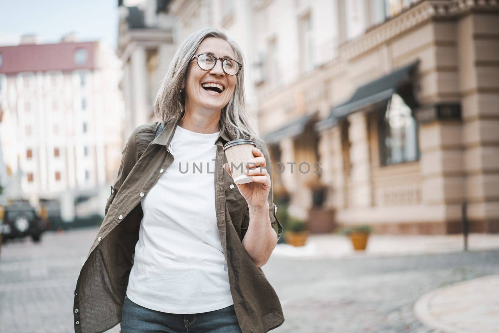 Concept of happy old age through image of silver-haired mature senior woman walking in city, enjoying cup of coffee. Woman is captured in moment of joy and contentment, with smile and positive mood. . High quality photo