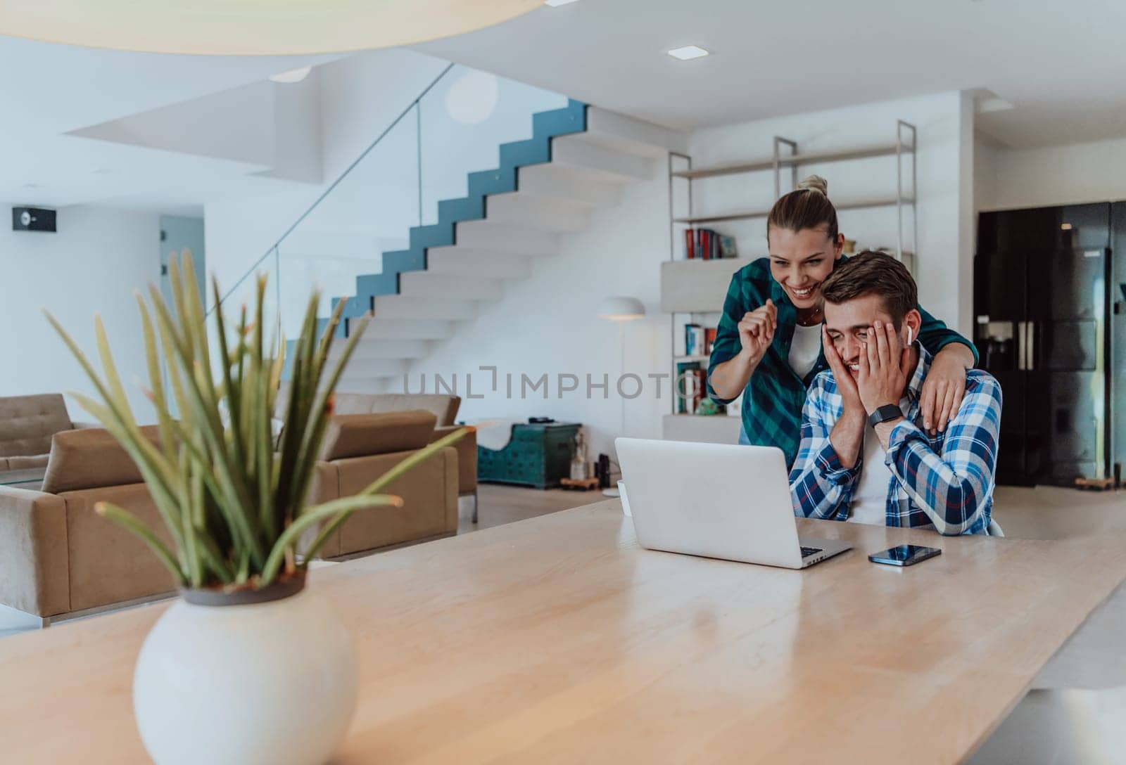 A young married couple is talking to parents, family and friends on a video call via a laptop while sitting in the living room of their modern house.