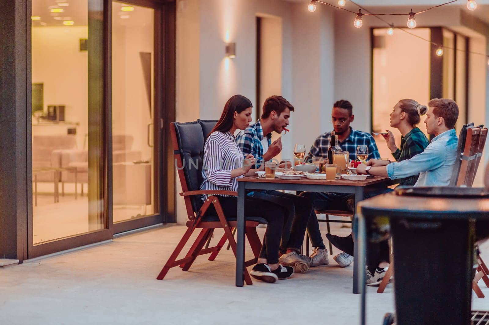 A group of young diverse people having dinner on the terrace of a modern house in the evening. Fun for friends and family. Celebration of holidays, weddings with barbecue. by dotshock